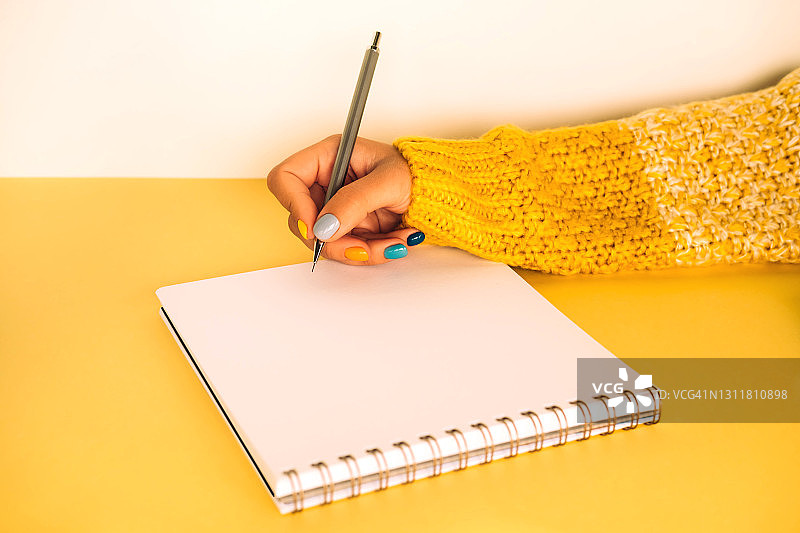 一个穿着黄白色针织毛衣，修着鲜艳多色指甲的女人正坐在黄色的桌子前写笔记本。2021年的流行色图片素材