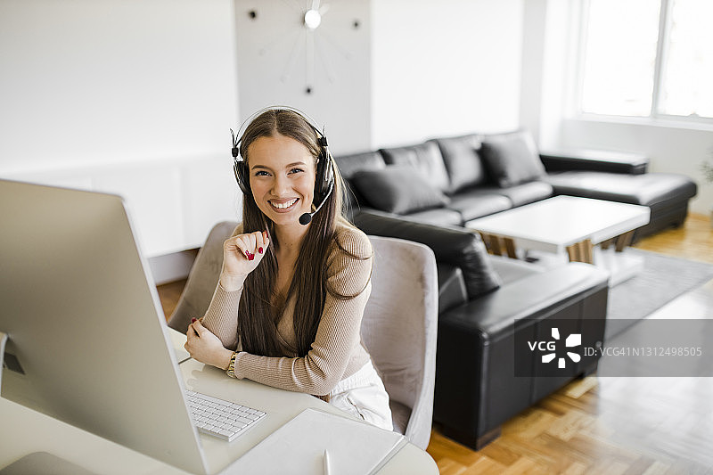 快乐的女人戴着耳机有视频电话和工作在她的家庭办公室的电脑图片素材