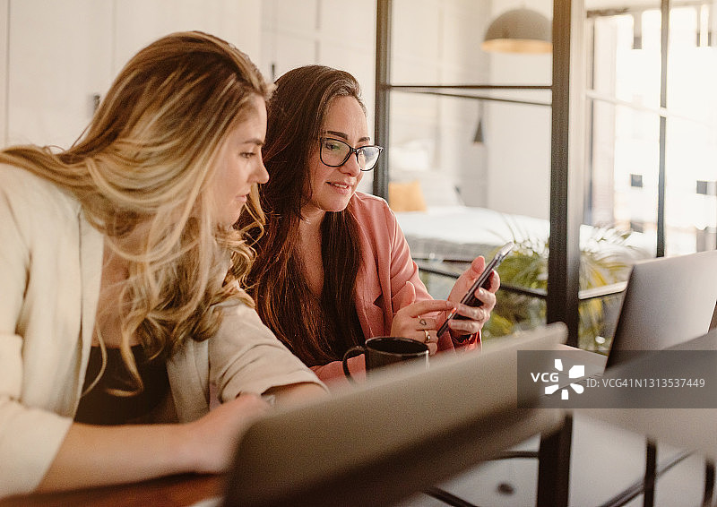 两个年轻的女商人在家工作，看着手机。营销战略头脑风暴。团队合作使女性。图片素材