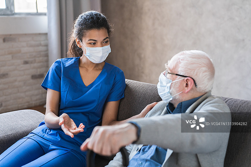 医生或女性护工，老人祖父戴着防护口罩在家或养老院保护自己免受Covid - 19感染。医生在家给老年病人谈话图片素材