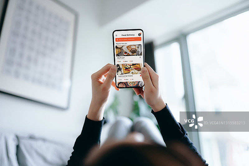 一名年轻的亚洲女性躺在舒适的家中的床上，用智能手机上的移动应用程序使用送餐服务和在线订餐。生活方式和日常生活中的科技图片素材