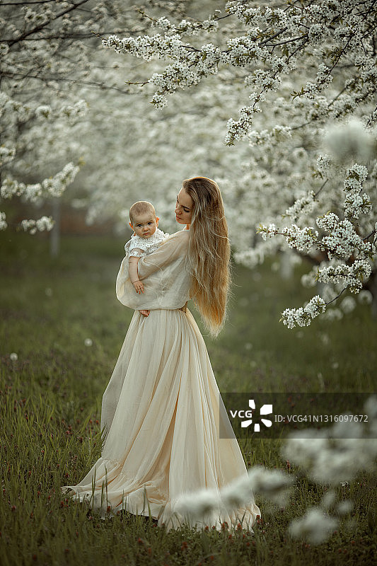 美丽的年轻母亲和婴儿女孩拥抱在花园的白色樱花在自然的夏天。夏日家庭度假。图片素材