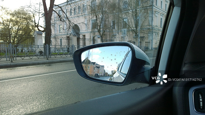 城市街道和天空在汽车侧镜与雨滴反射。汽车在雨中行驶。透过车窗的城市景观。乘坐汽车。真实的生活。城市环境。公路旅行。图片素材