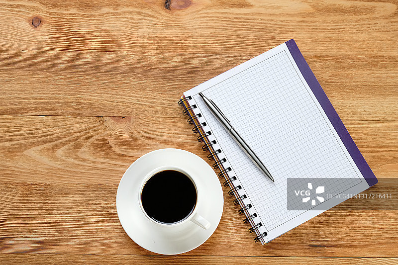一支笔在一张空白的记事本和一杯黑咖啡在木制餐桌上。商人或经理在工作场所或桌面上的物品。咖啡休息时间或商务会议，制定一天的计划。文本空间的拷贝。图片素材