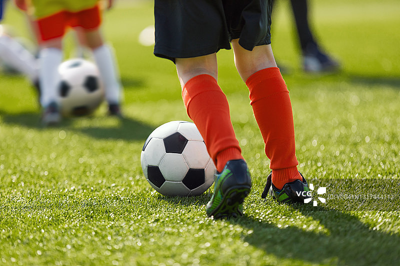 儿童户外足球训练。儿童足球运动员在草地上踢球的腿。儿童足球暑期学校。身穿彩色运动制服及足球钉鞋的青年运动员图片素材