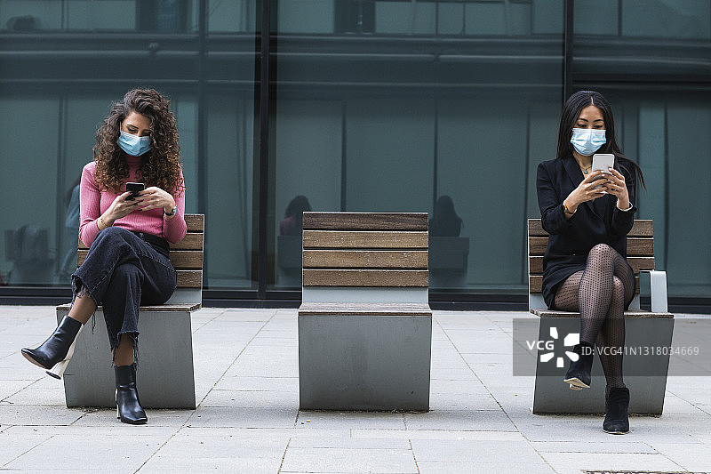 年轻女性戴着保护面罩使用手机，坐在长椅上保持社交距离图片素材