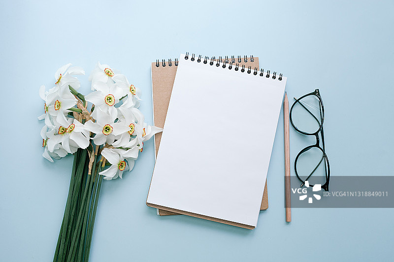 一束春天的白色水仙花，空白白纸的便条本，眼镜和铅笔在蓝色的背景。春节贺卡，母亲节。图片素材