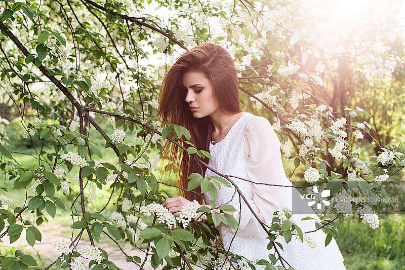一个年轻的黑发女孩穿着白色短裙，在白色的鸟樱桃树附近摆姿势，笑着享受大自然图片素材