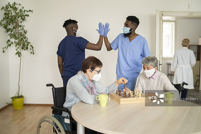 两名年长的白人女性坐在轮椅上，戴着防护口罩下棋，两名同样戴着防护口罩和手套的男性护士在后面击掌图片素材