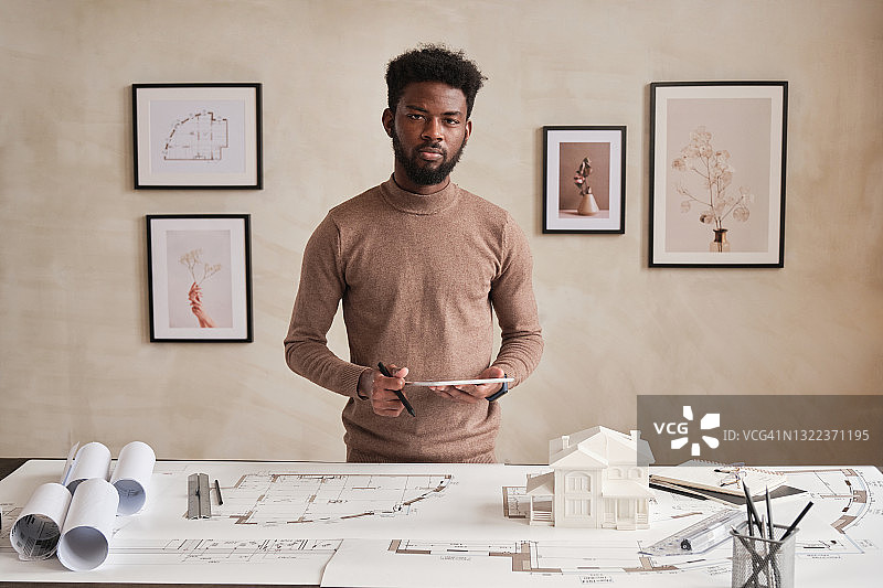 肖像严肃的年轻留胡子的黑色建筑师站在毛衣与写字板桌旁满是草图和当代办公室用品图片素材