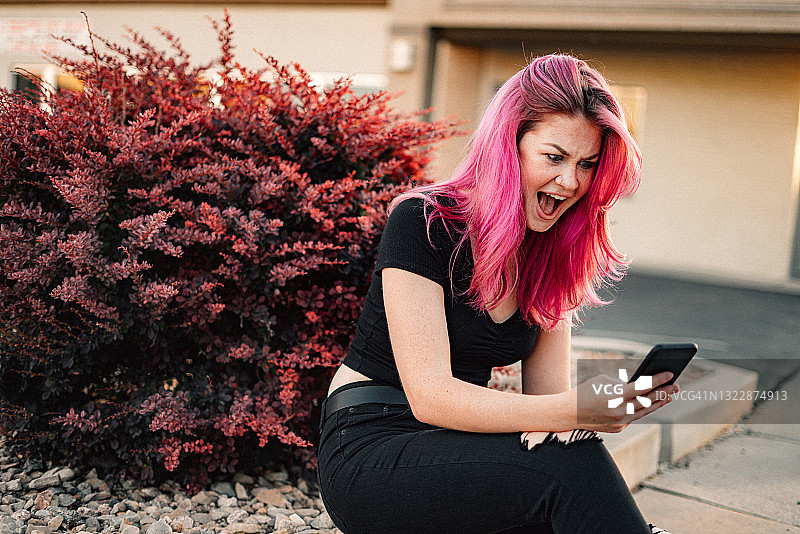 一个时尚的年轻女人的肖像与有趣可爱的亮粉色染头发用她的智能手机和在夏天户外做一个脸图片素材