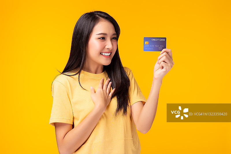 年轻的亚洲女人展示塑料信用卡和看着信用卡与兴奋和快乐站在黄色的背景美丽的女孩得到满意的信用卡服务与微笑的脸图片素材