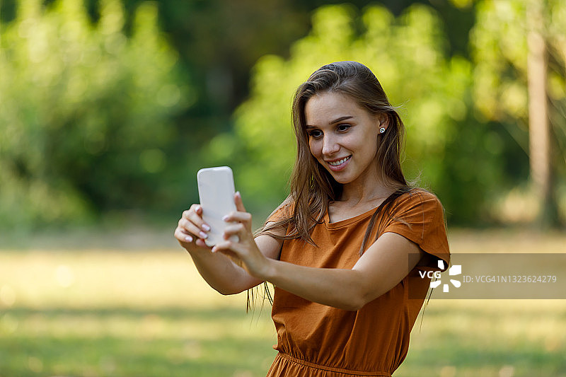 一位年轻女子在公园里散步时，正在用手机自拍。图片素材