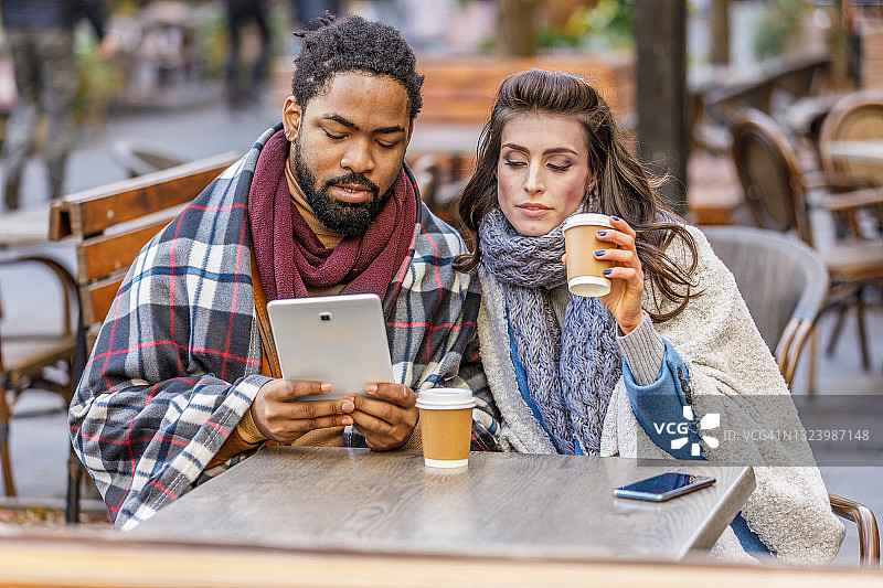 一个年轻的非洲男人和他的妻子正坐在户外咖啡馆的桌子旁，一边喝着咖啡，一边使用着数字平板电脑。图片素材