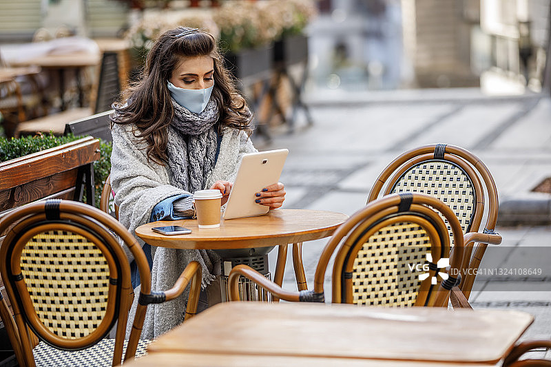 美丽的女人戴着面具坐在餐厅喝咖啡和上网。图片素材