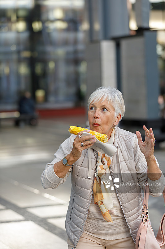 一位老妇人在城市里散步，享受玉米芯。图片素材