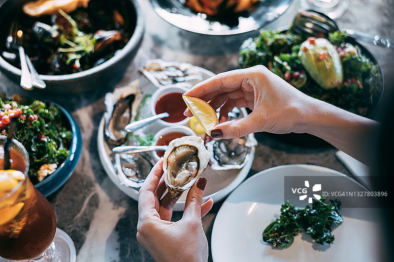在餐馆里，一位女士正在享用美味的一餐，她的手正在往一只新鲜的牡蛎上挤柠檬汁。外出就餐的生活方式图片素材