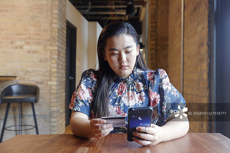 一位留着长发的亚洲千禧一代女性穿着休闲装，在家里用手机刷信用卡付款。图片素材