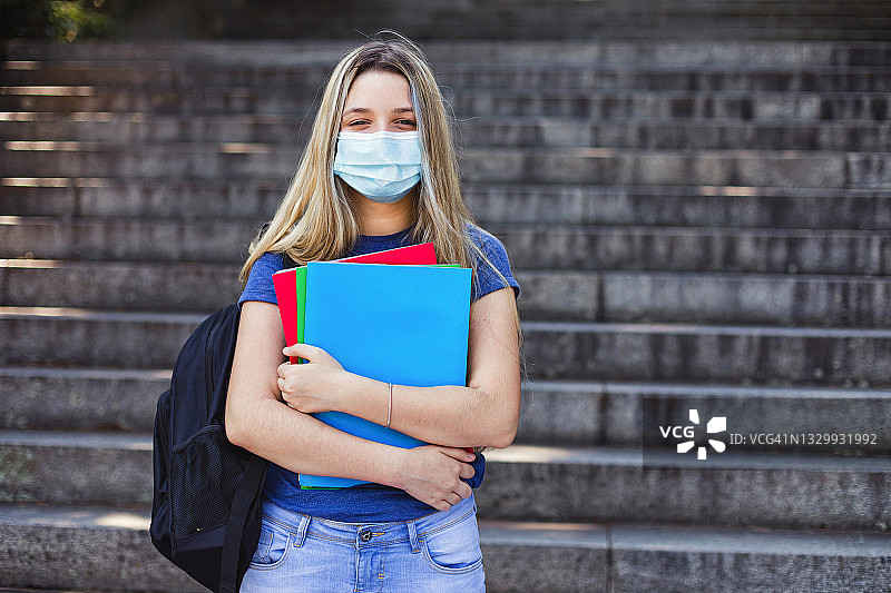 女学生在学校里拿着书走。她戴着口罩来保护新冠病毒。回到学校的概念图片素材