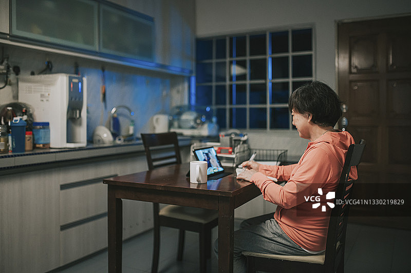 一名亚洲华裔老妇人晚上在厨房用数字平板电脑与她的儿子和儿媳进行视频通话图片素材