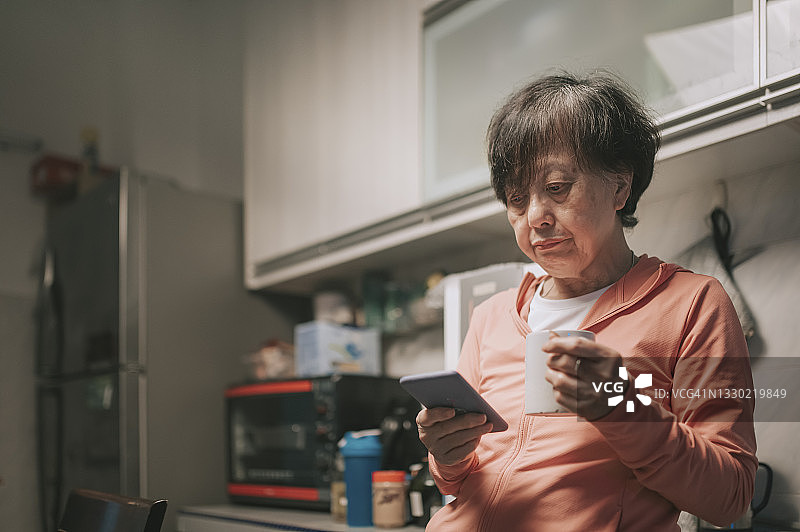 晚上，亚洲华裔老年妇女在家庭厨房里倚靠着手机，喝着咖啡图片素材