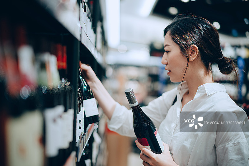 年轻的亚洲女人走过超市的酒类通道，从货架上挑选一瓶红酒图片素材