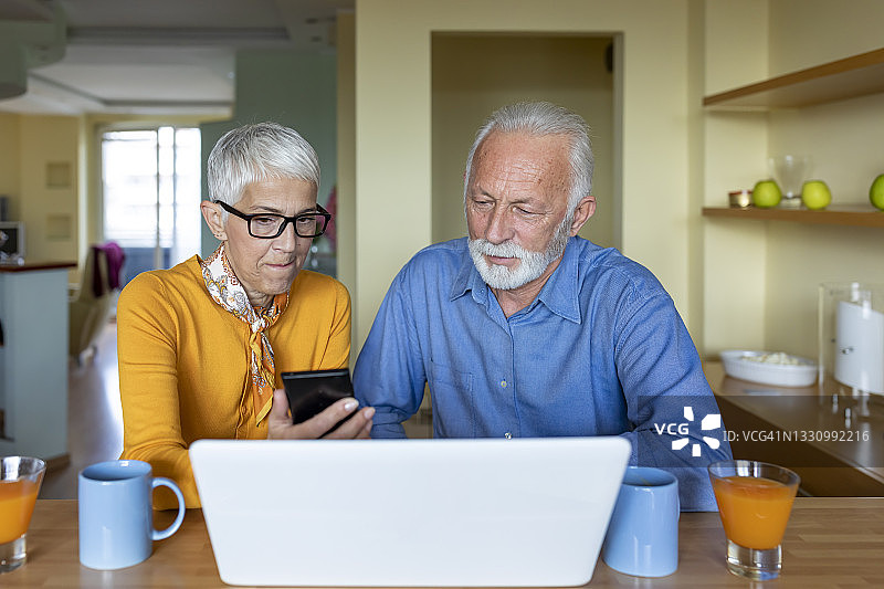 一位高级男士和他的妻子坐在家里的桌子旁，一边喝着咖啡，一边用笔记本电脑和电话上网。图片素材