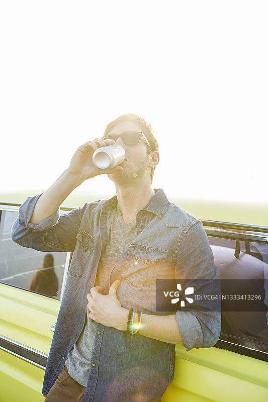 年轻人靠在汽车上，靠在晴朗的天空，喝着啤酒图片素材
