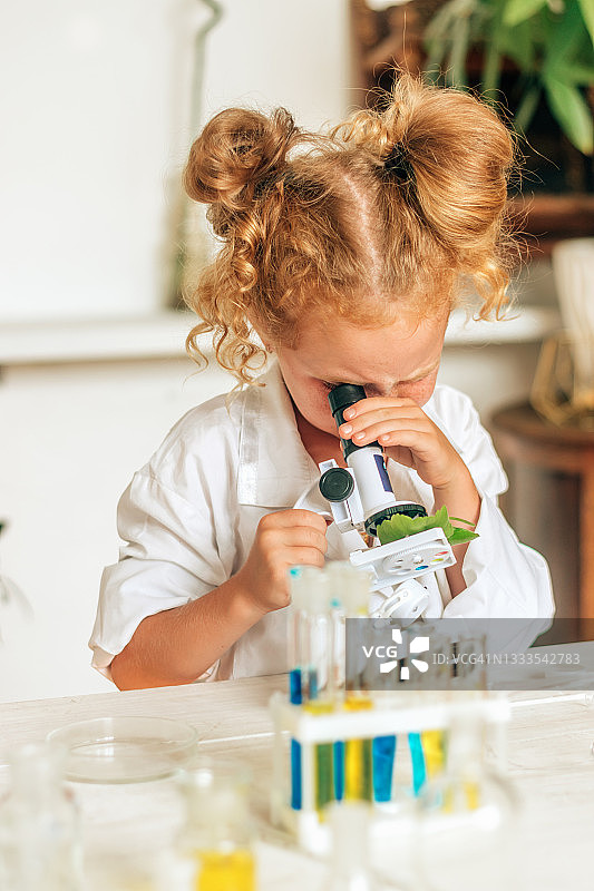 小女孩穿着白色制服，戴着防护眼镜，在一个家庭实验室里看着显微镜。回到学校的概念。年轻的科学家。自然科学。儿童的学前和学校教育。图片素材