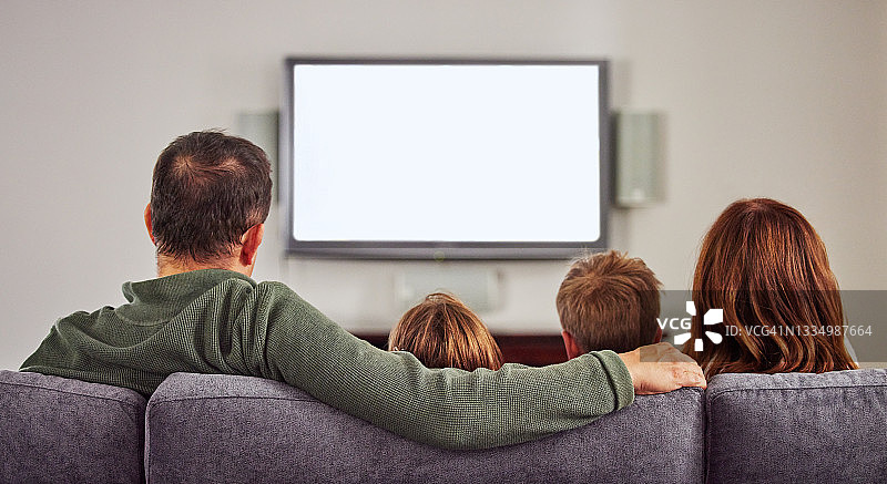 照片中，一个陌生的家庭坐在家里的沙发上，聚在一起看电视图片素材