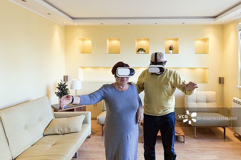 快乐的老年妇女和她的丈夫在家中客厅的虚拟现实中享受着手势。图片素材
