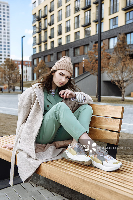 一个戴着帽子、穿着大衣的年轻女子坐在城市街道背景下的一条长凳上图片素材
