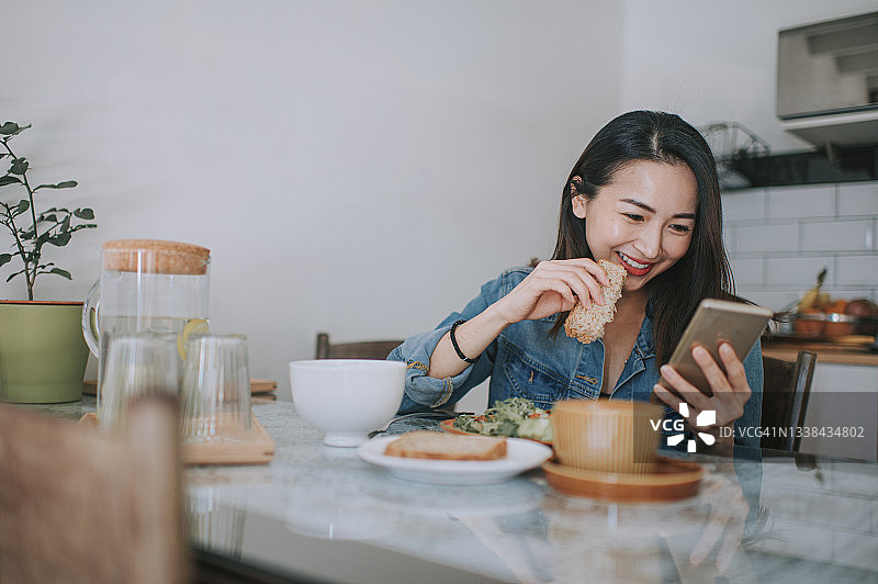 亚洲华裔美女在她的智能手机上阅读，在家里享受她的早餐，用全麦面包涂花生酱和咖啡图片素材