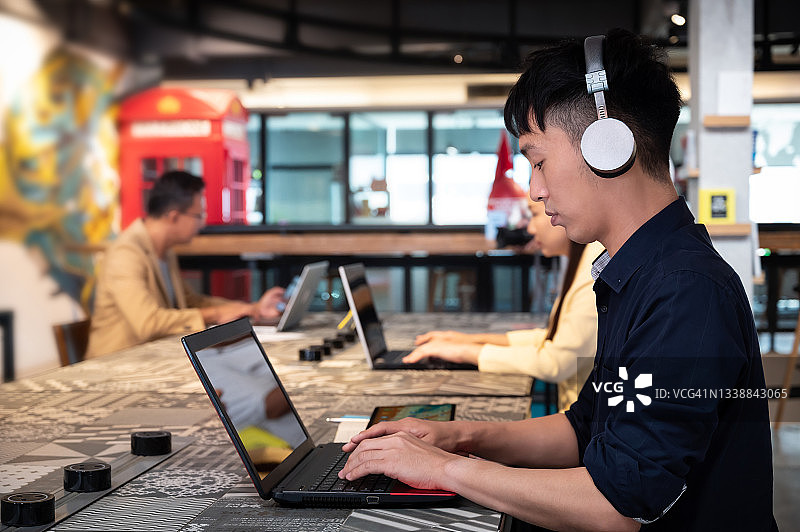 年轻的亚洲人听音乐与无线耳机和工作在共同工作的办公室。图片素材