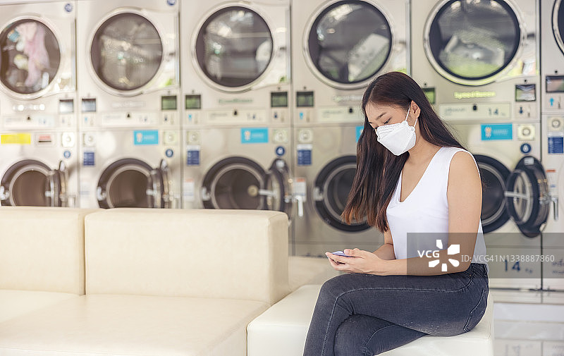 在一个有很多自动洗衣机和模糊背景的自助洗衣店里，一位亚洲女性坐在那里，用她的智能手机做着自由职业工作。图片素材