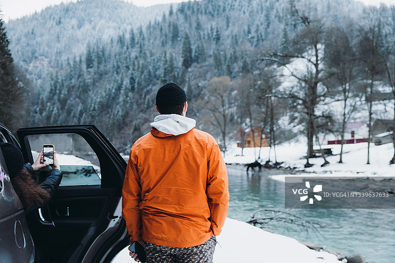 穿着橙色夹克的年轻男子和拿着智能手机的女子在靠近汽车的山上享受冬天图片素材