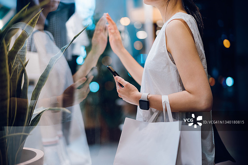 黄昏时分，一名年轻的亚洲女子背着一个纸质购物袋，站在一家精品店外看橱窗，查看自己的智能手机图片素材