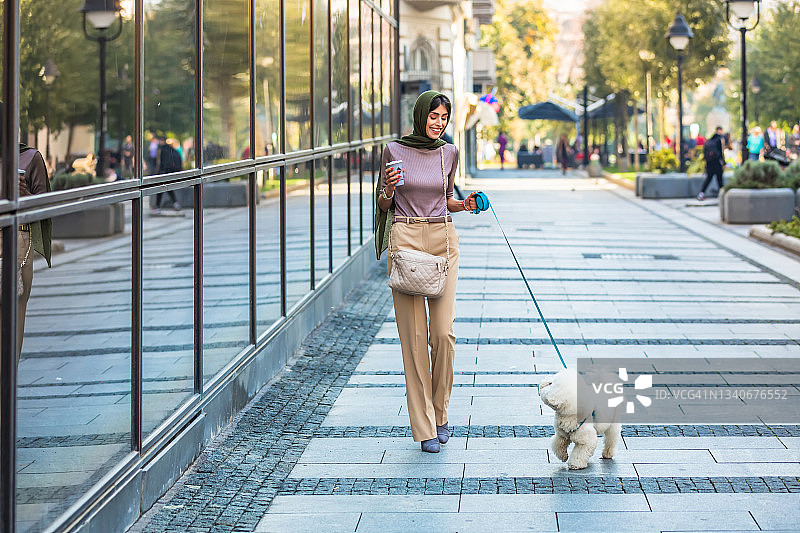 背景中，一名穆斯林妇女穿着暖和的衣服在办公楼附近遛着她的小狗。图片素材