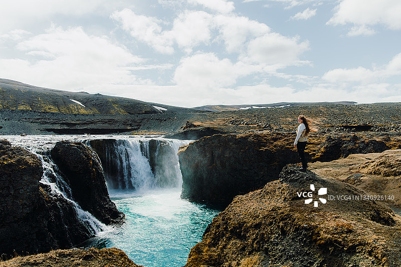 一个女人在阳光明媚的刮风的日子里从冰岛的山顶上看风景优美的瀑布图片素材