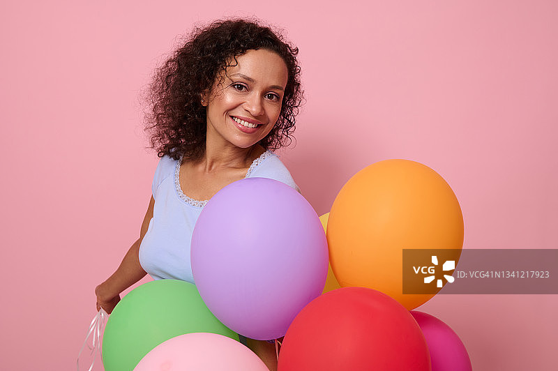 迷人的美丽的混血儿女人带着迷人的牙齿微笑，抱着鲜艳的彩色氦气球有快乐的反应，庆祝生日，看着相机，孤立在粉红色的背景上图片素材