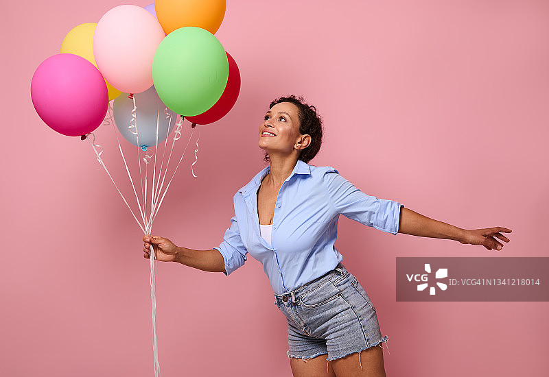 快乐微笑的年轻混血非洲裔美国妇女与一束彩色彩虹气球向上看，并有乐趣，穿着牛仔短裤和蓝色衬衫在粉红色的墙背景与复制空间图片素材