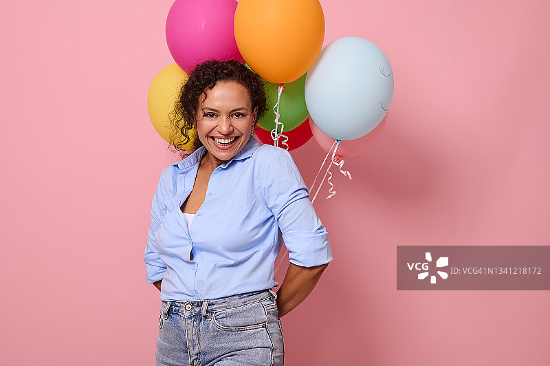 可爱快乐的非洲年轻女子身穿蓝色衬衫，微笑着看着镜头，背后拿着五颜六色的气球。粉红色背景，复制空间图片素材
