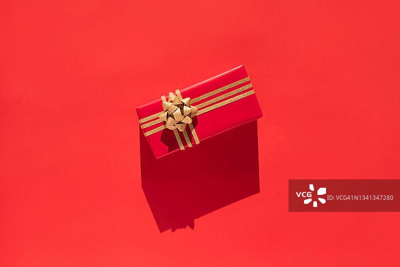 红色礼品盒与金光闪闪的弓在红色背景与阴影。平躺风格图片素材