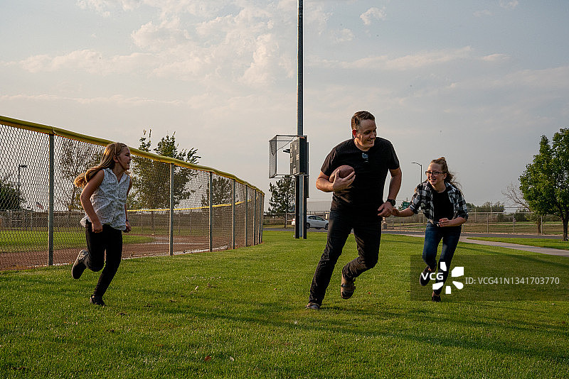 年轻的父亲和他的两个女儿在公园的草地上玩触身式足球图片素材