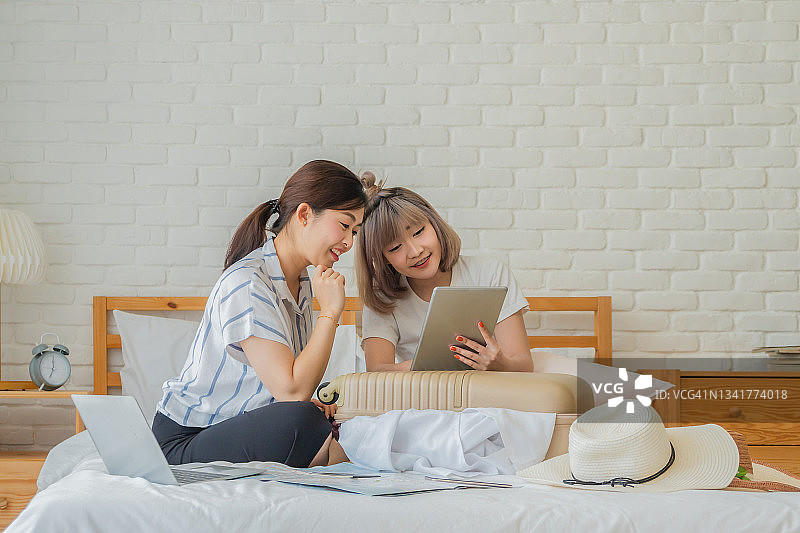 两个年轻的亚洲女性朋友在家里的床上用平板电脑为旅行旅行准备和打包衣物图片素材