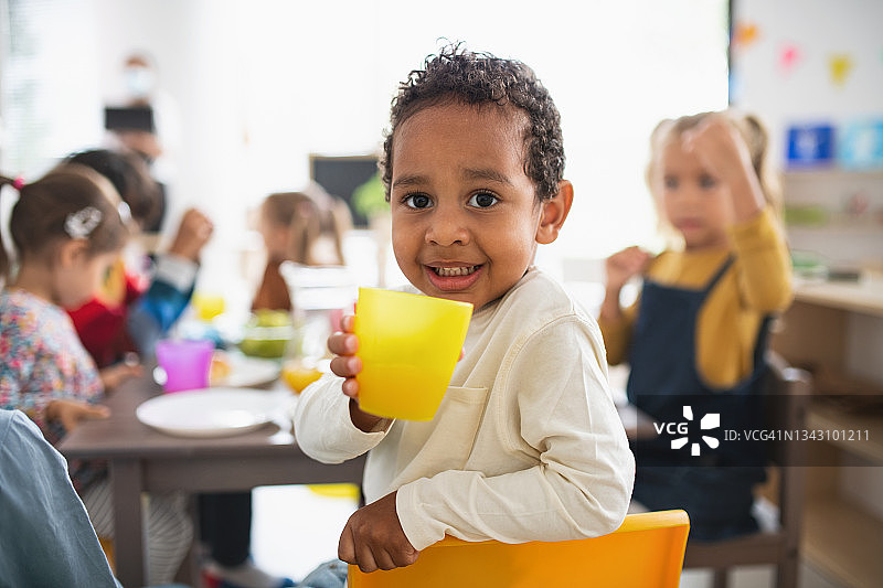 学龄前多种族男孩在幼儿园与孩子们在室内吃饭，看着相机。图片素材