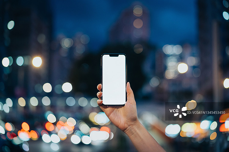 一名女子手持智能手机，背对着灯火通明的市中心街道，以夜晚繁忙的城市交通为背景。智能手机与空白屏幕的设计模型图片素材