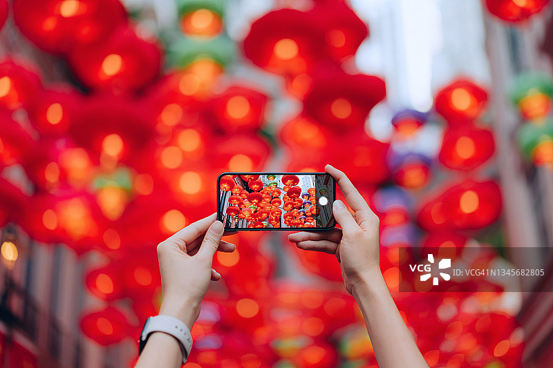 一名女子手拿智能手机在城市街道上为中国传统红灯笼拍照。中国传统文化、节日和庆典活动主题图片素材