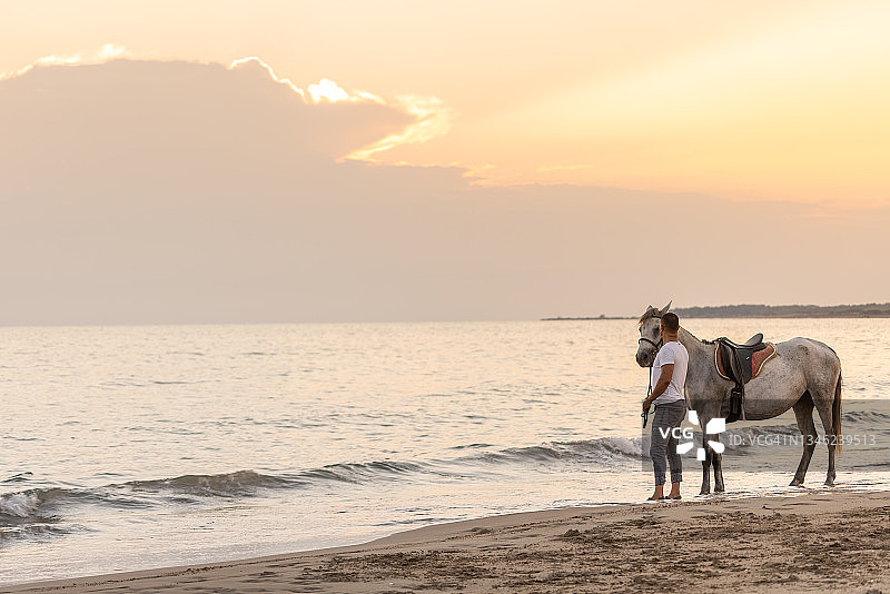 一个男人和他的马站在日落的海滩上。图片素材