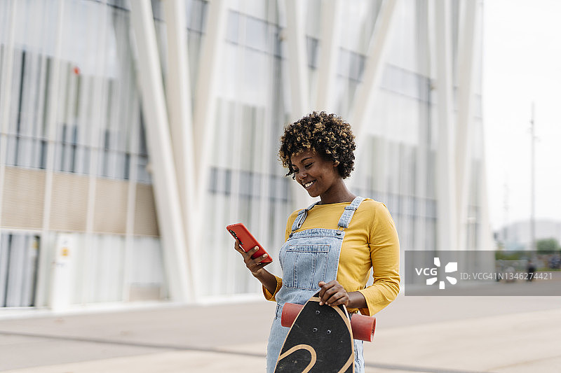 一个穿着非洲式滑板的女人微笑着在街上玩手机。图片素材